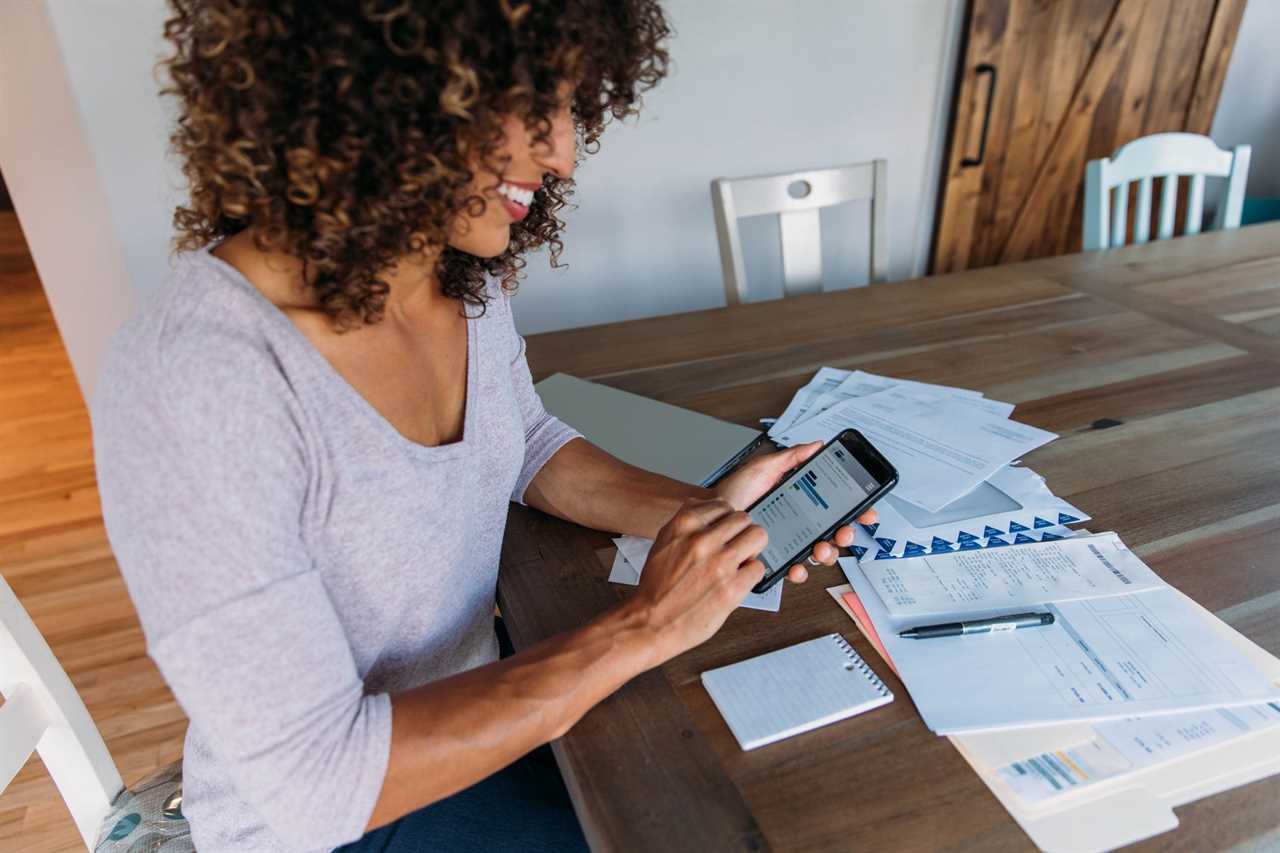 woman sitting in kitchen table looks over finances and uses phone to track spending and budgeting