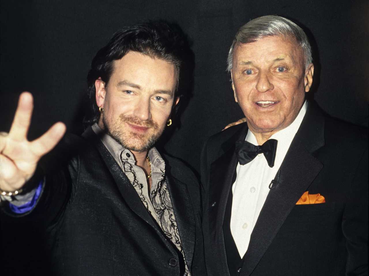 Bono giving a peace sign and embracing an older Sinatra who is smiling in a tuxedo.