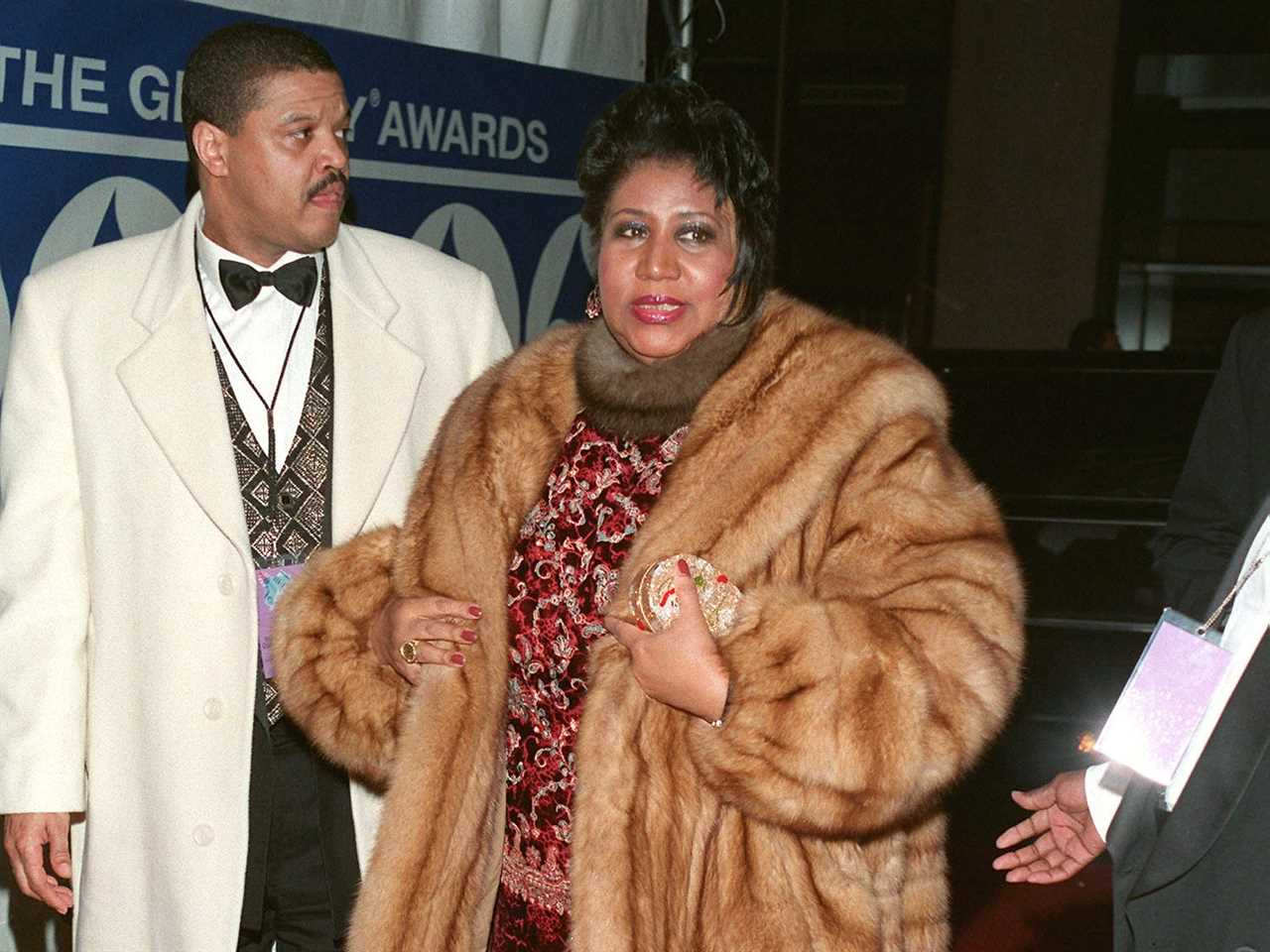 Aretha in a burgundy gown with black lace and gold embellishments and a brown fur coat and scarf walking on the red carpet.