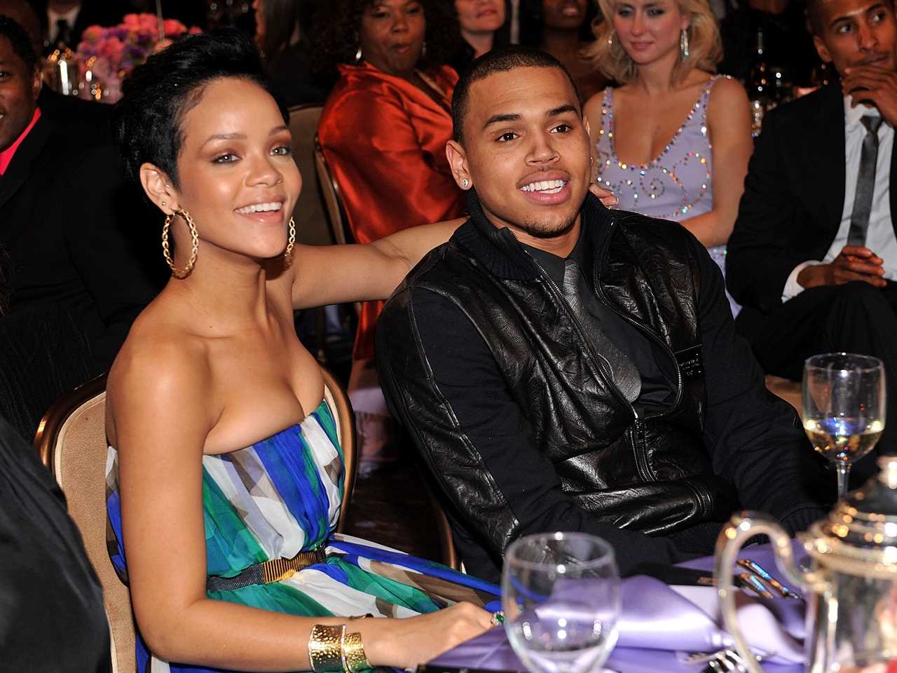 Rihanna in blue, green, and white striped strapless dress with her arm around Chris in a black leather jacket. Both are smiling and sitting at a dinner table.