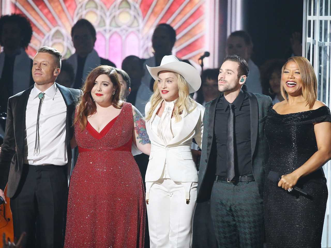 Macklemore, Mary Lambert, Madonna, Ryan Lewis, and Queen Latifah hugging and smiling in front of a choir and a stained-glass window design.