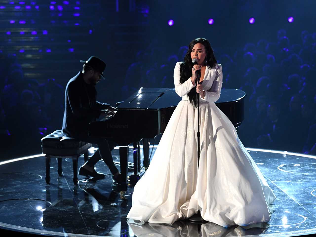 Demi in a large white ballgown singing into a microphone on stage with a piano player performing behind her.