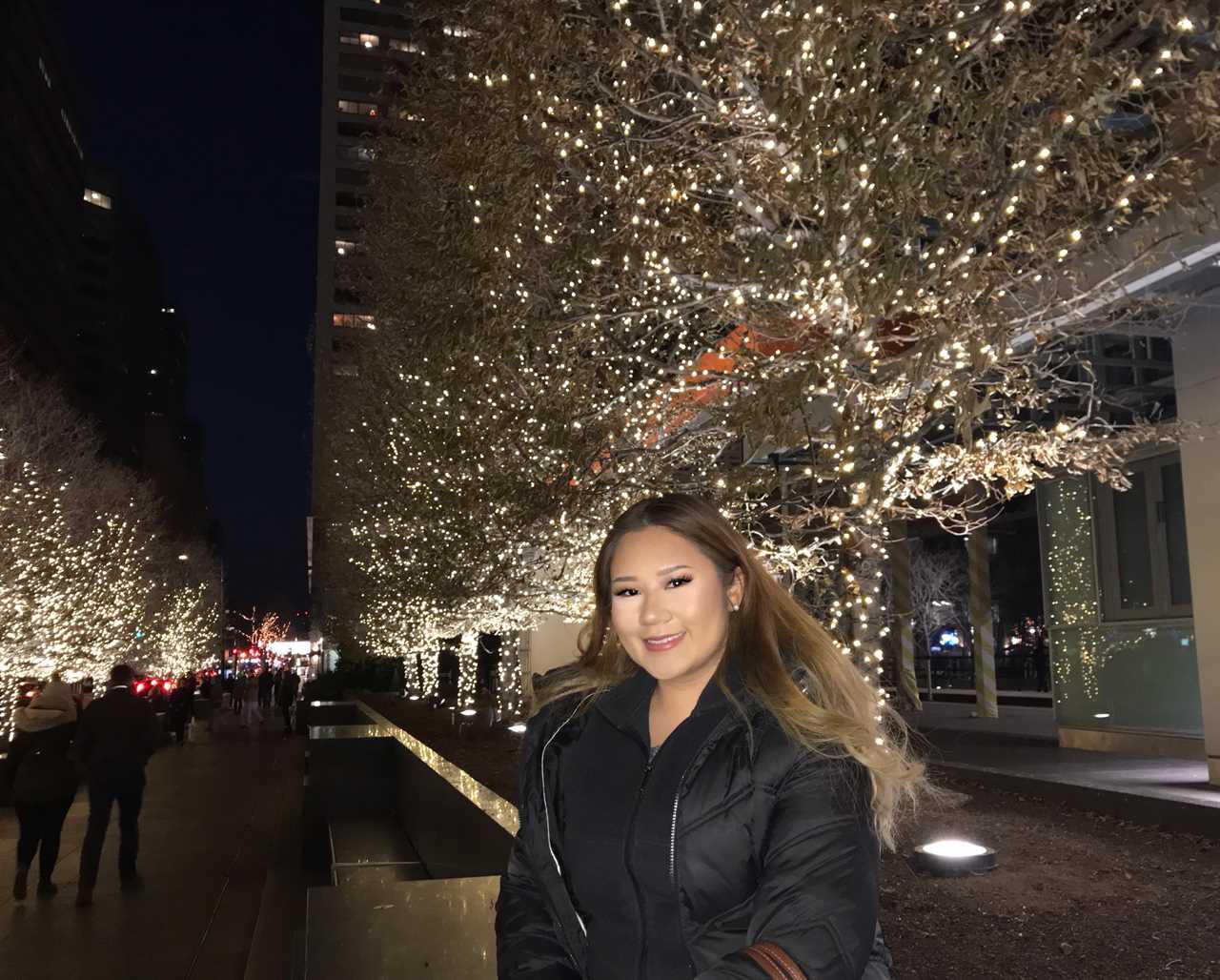 Kelsey Lam wearing a black coat and standing in front of trees with lightbulbs strewn around the branches.