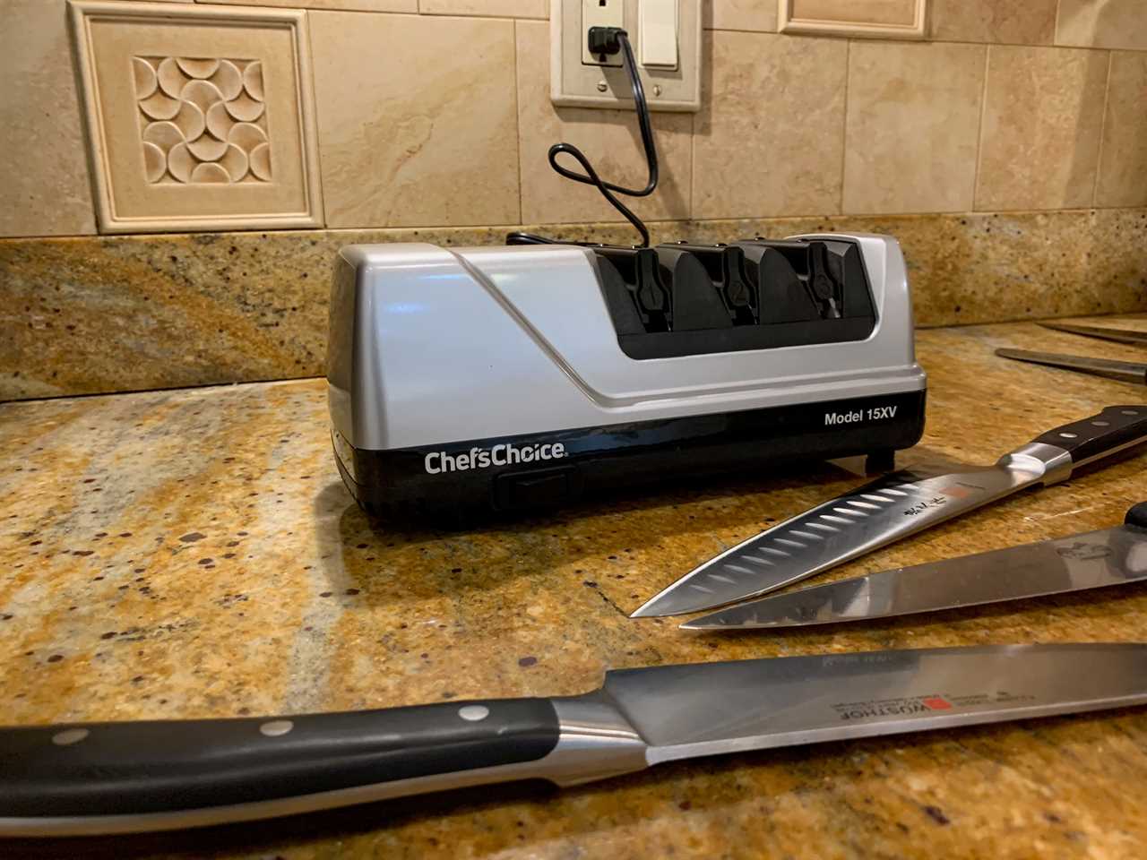 Several chef’s knives laid out on a granite countertop in front of the Chef’s Choice Trizor XV EdgeSelect knife sharpener.