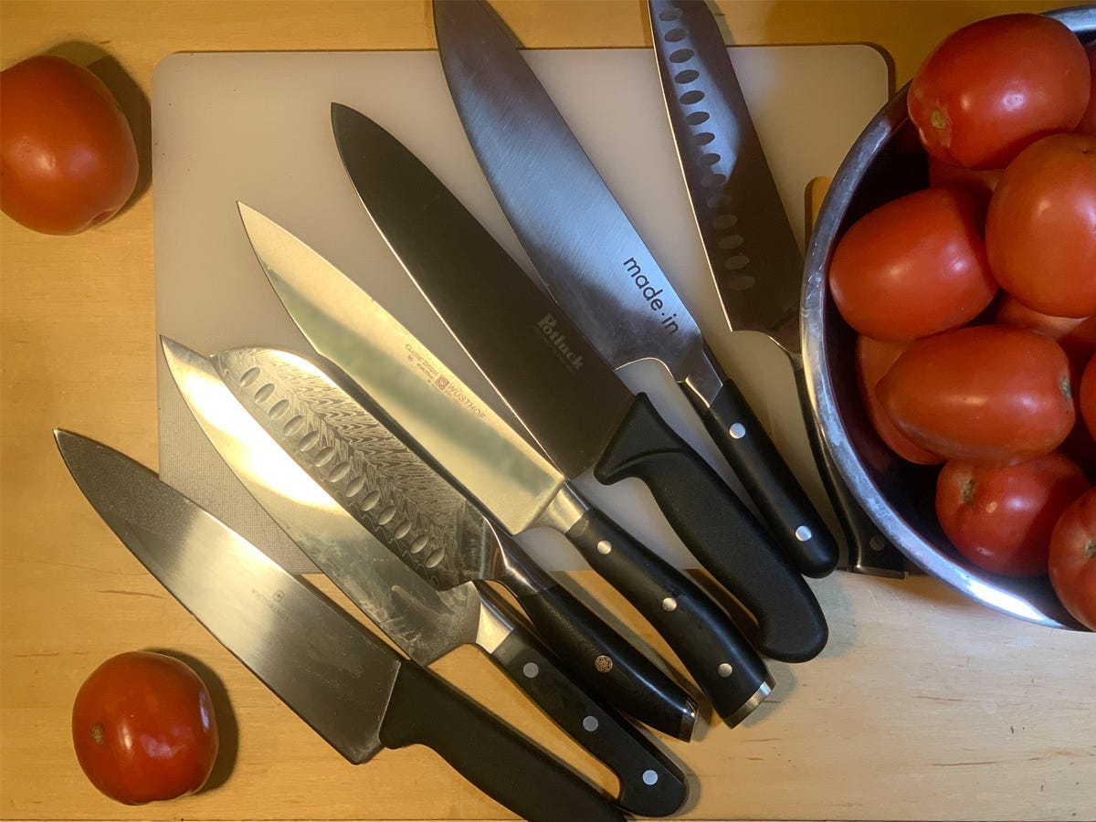 Several chef's knives laid on top of a cutting board beside a bowl of tomatoes.