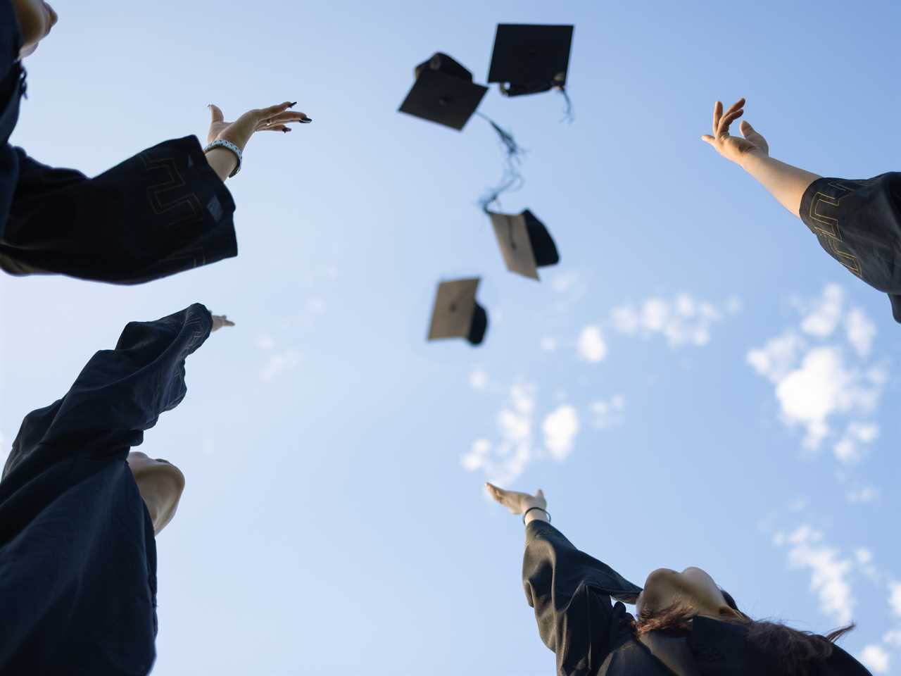 College graduates in gowns through their hats up toward a blue sky.