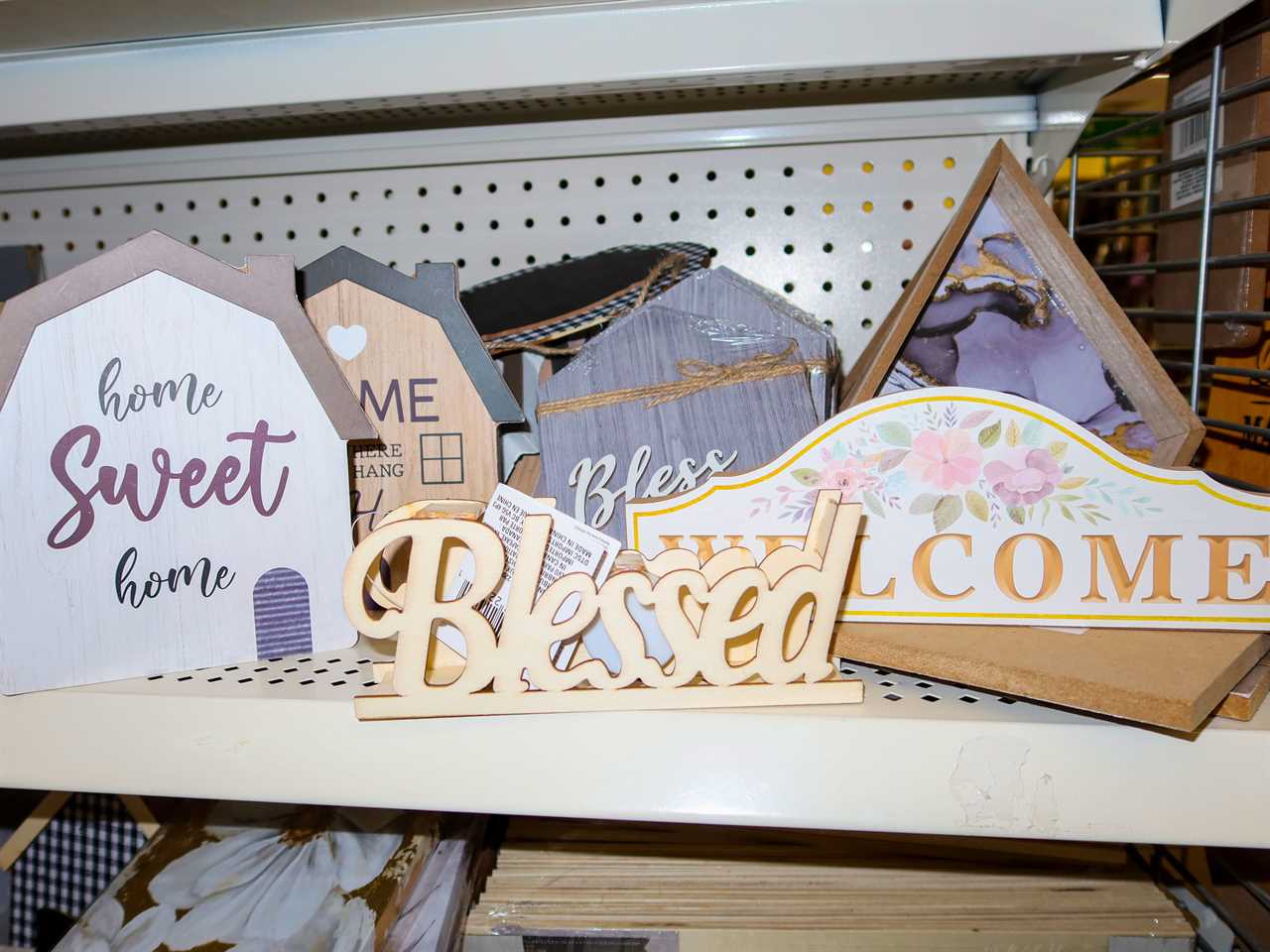 Shelf of Decor with short sayings on them at Dollar Tree. Signs include a house silhouette with "home sweet home," a "welcome" plaque, and a "blessed" sign