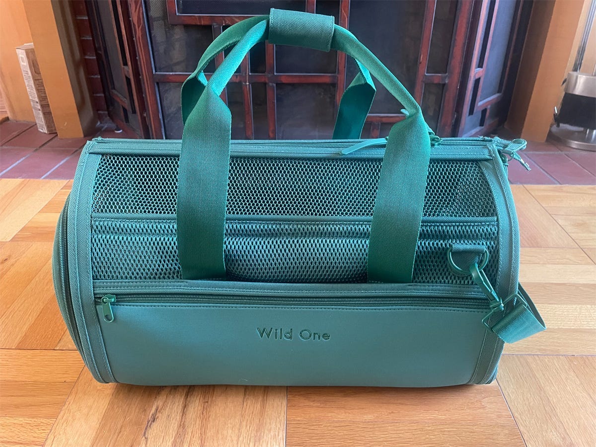 A teal soft-sided airline approved pet carrier with a mesh top set on a wooden floor.