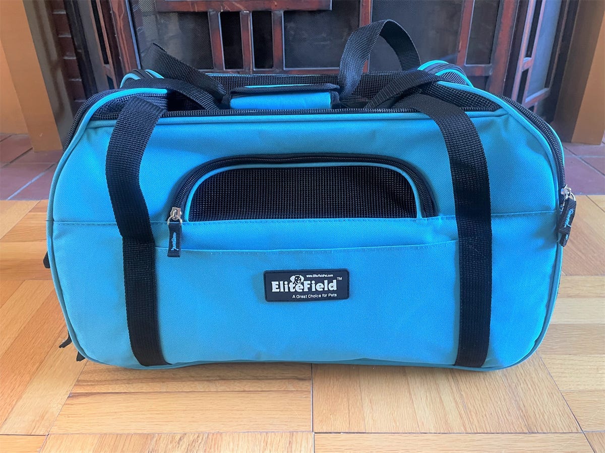 A soft-sided blue cloth Elite Field Soft-Sided Airline Approved Carrier sitting on a wooden floor.