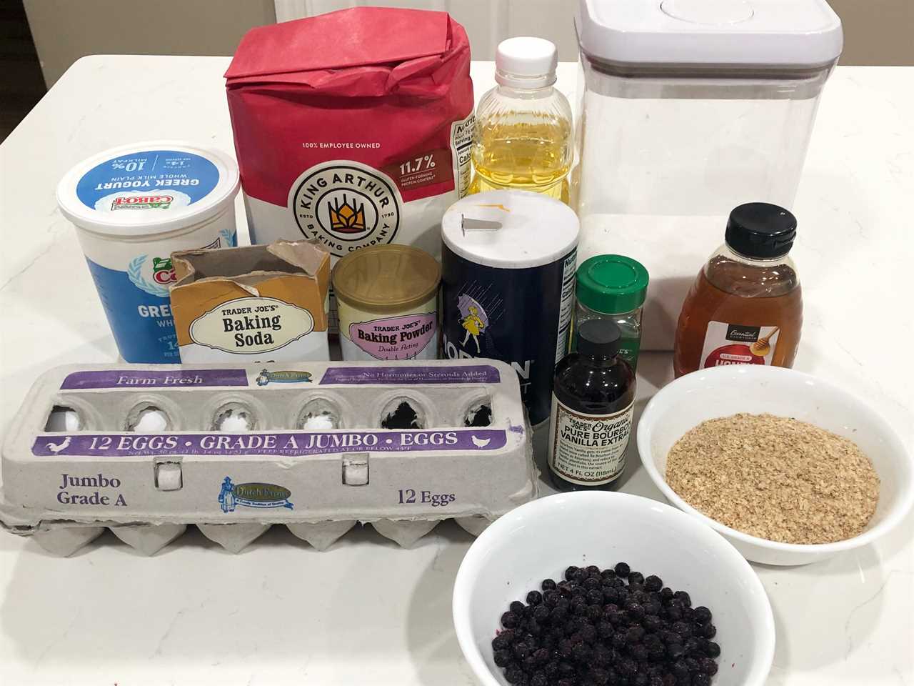 Containers of Greek yogurt, baking soda, baking powder, salt, flour, sugar, vegetable oil, vanilla extract, honey, and cinnamon, a carton of eggs, and bowls of wheat bran and blueberries.