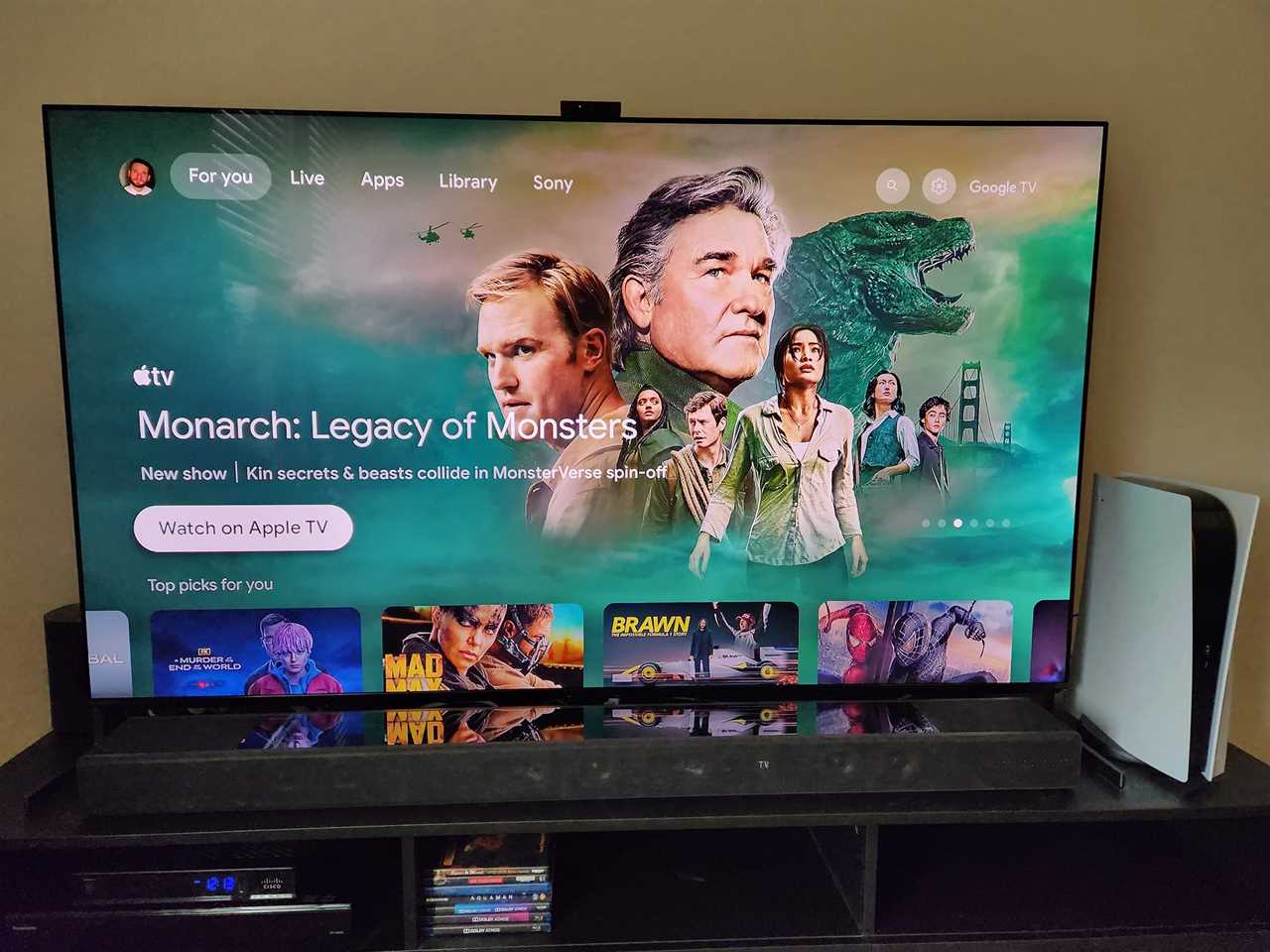 A front view of a Sony A95L 4K OLED TV on a TV stand displaying the Google TV home page with an image from "Monarch: Legacy of Monsters" on the screen.