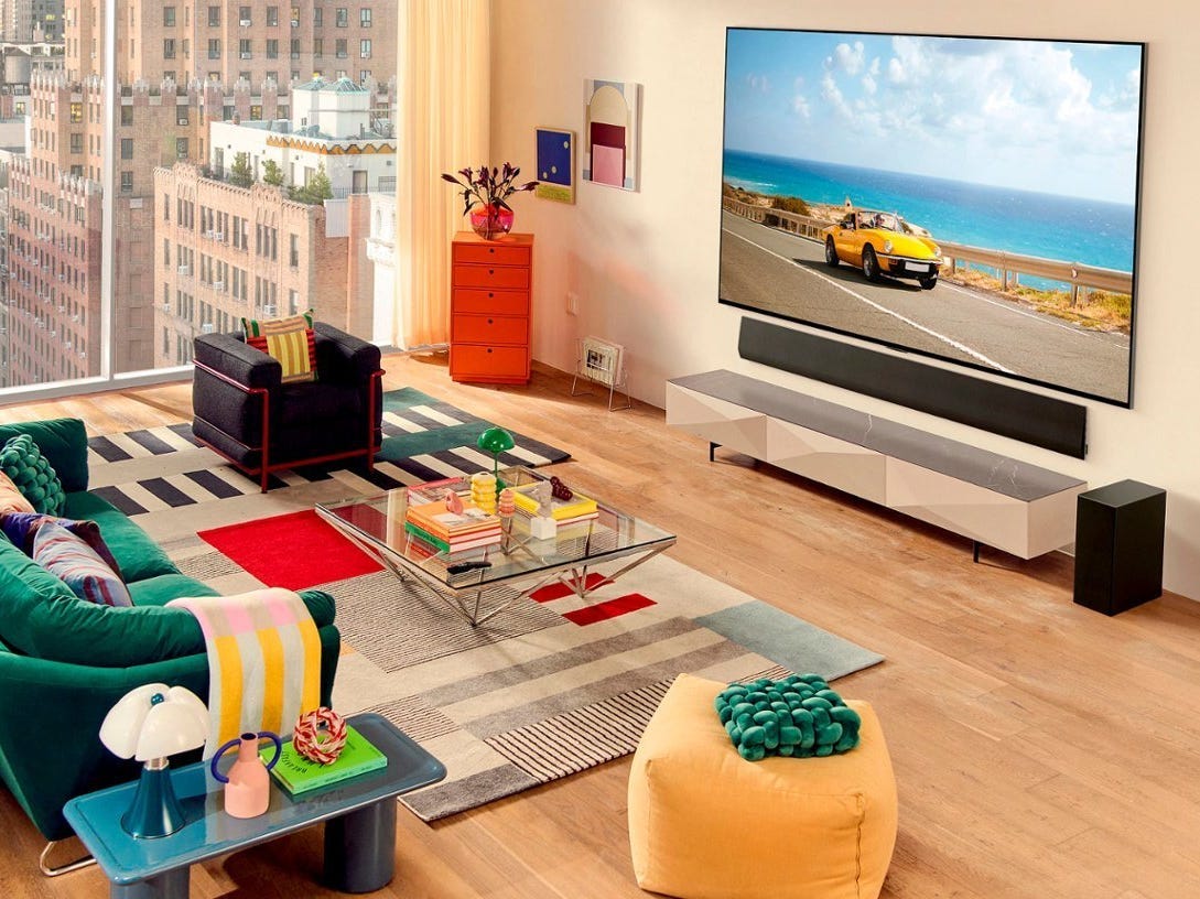 An LG G3 4K OLED TV hanging on a wall above a soundbar and TV stand across a green couch in a living room during the day.