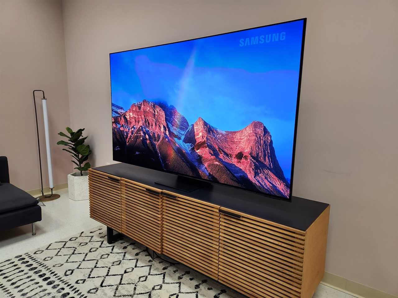 A mountain range displayed on the screen of a Samsung S95C TV that's on top of a media stand in a bright room.