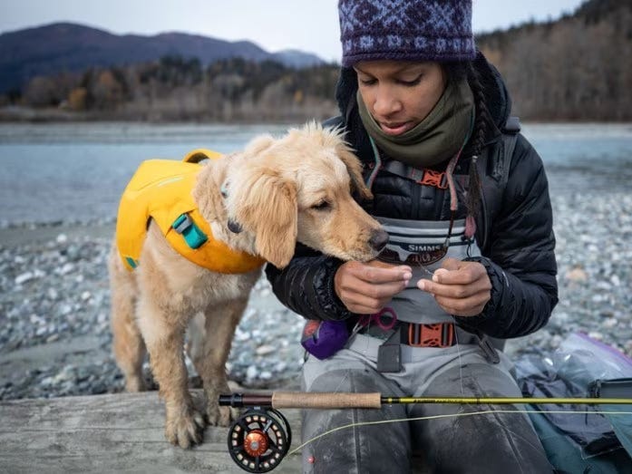 A yellow lab dog is standing on a rocky beach beside a person with a fishing pole with the water behind them.