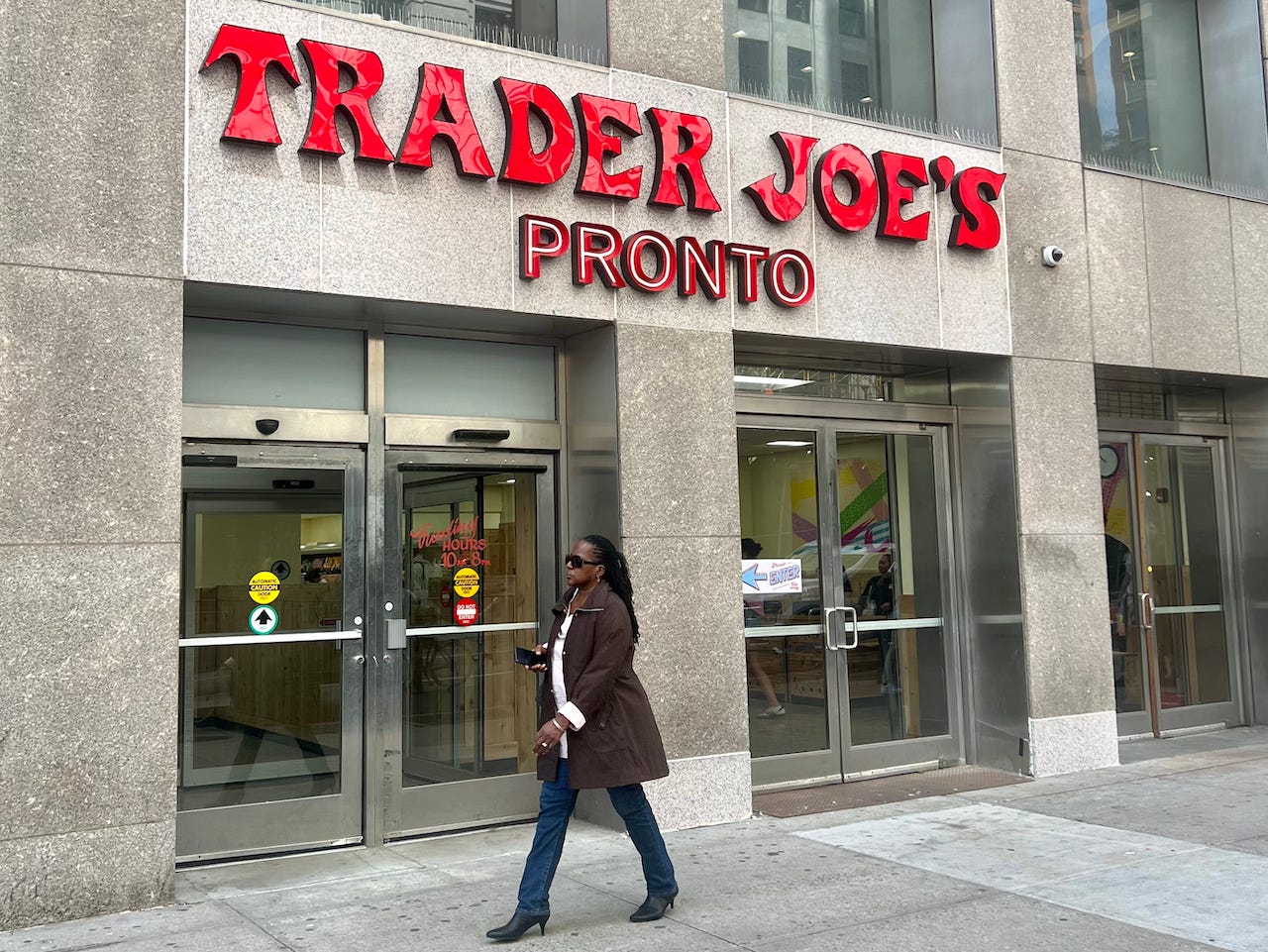 The entrance to the grab-and-go store with big red letters that say Trader Joe's Pronto on the gray facade over silver doors.