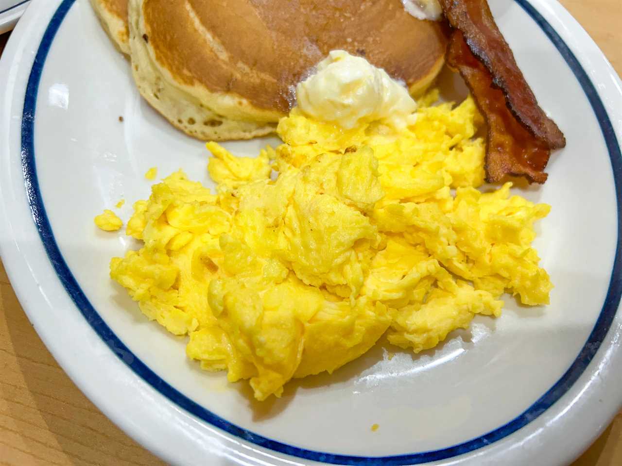 Scrambled eggs on a plate with a blue rim. Pancakes and bacon also sit on the plate