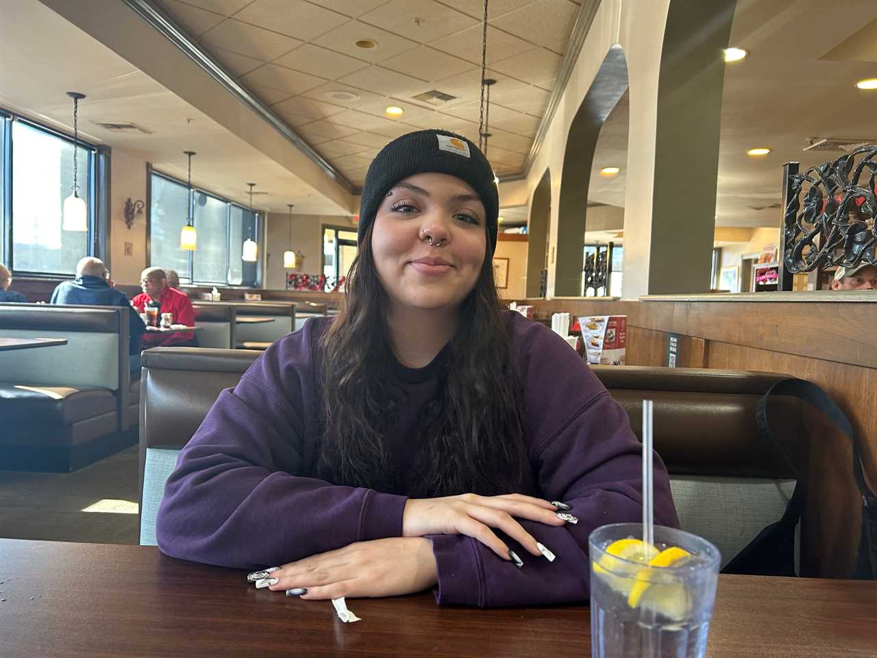 Picture of the writer wearing a purple sweatshirt and a black beanie in a booth at Perkins. People sit at other booths in the background