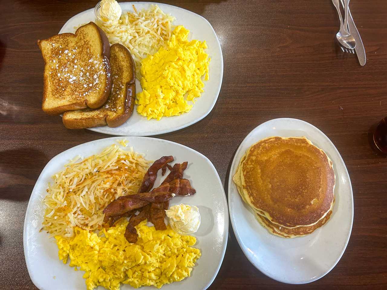 Three plates of breakfast food on a table at Perkins. Plates have French toast, hash browns, and scrambled eggs; hash browns, bacon, and scrambled eggs; pancakes