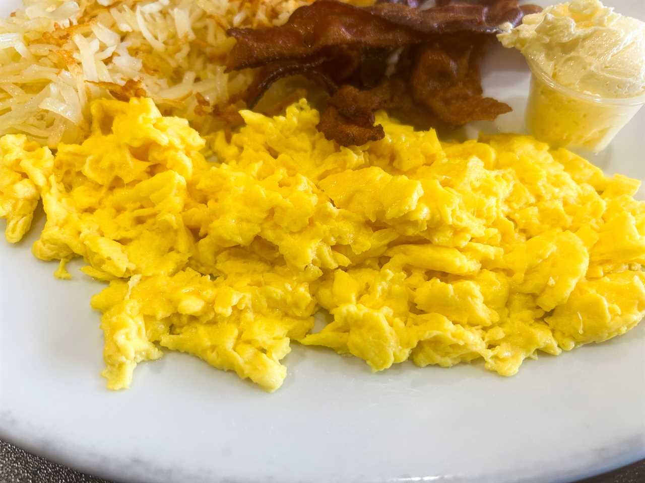 Scrambled eggs on a white plate with hash browns, bacon, and butter in the background