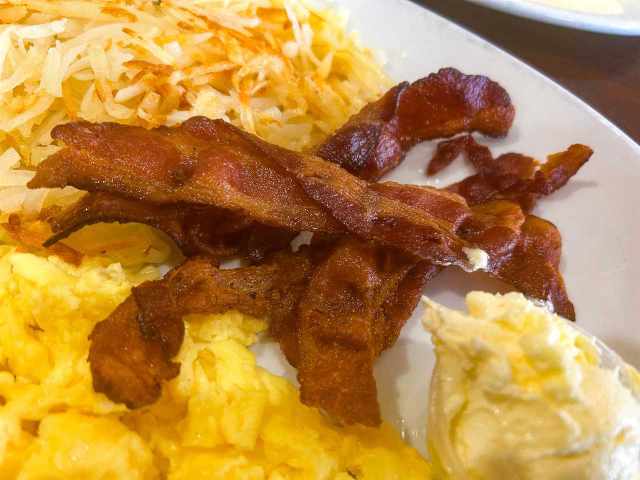 Plate of hash browns, butter, scrambled eggs, and bacon. The bacon looks crispy