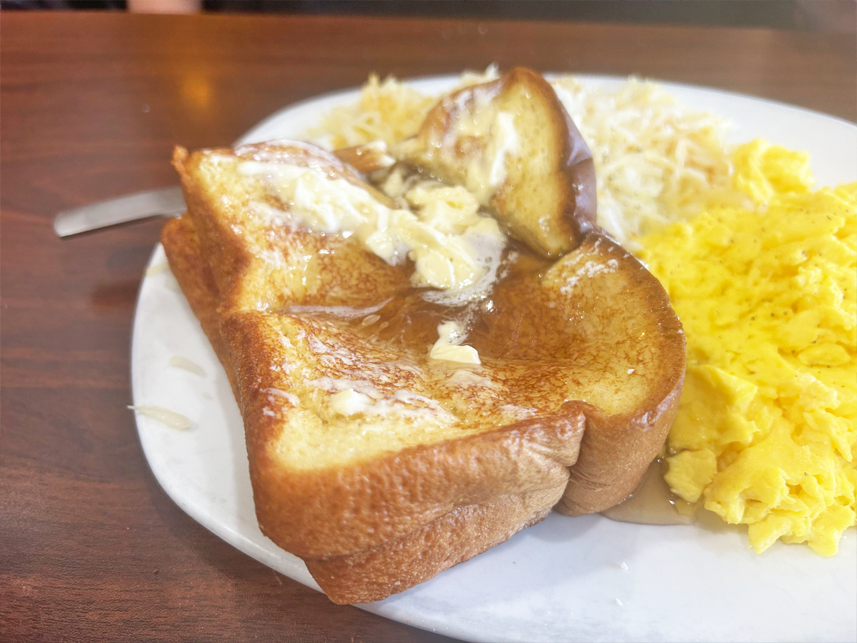 Two slices of French toast with syrup and butter on it. The French toast sits on a white platter with scrambled eggs and hash browns
