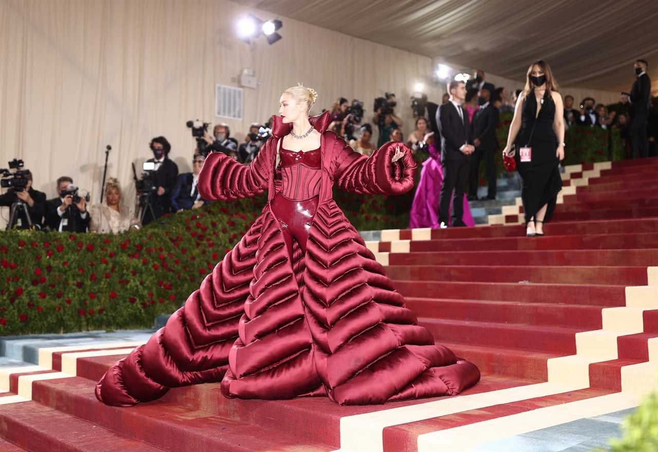 Gigi Hadid poses on the Met Gala red carpet on its famous stairs in a large red coat with a corset underneath.