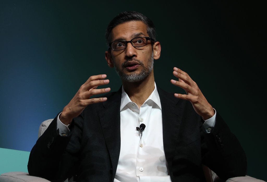 Google CEO Sundar Pichai sits on gray armchair and gestures with both hands while giving a speech.