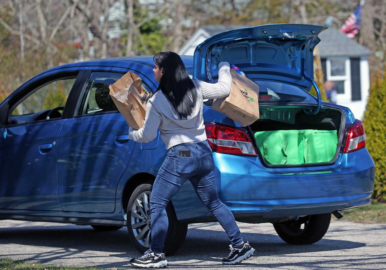 An Instacart shopper grabs paper bags full of groceries out of the back of a blue compact car while wearing a sweater, blue jeans, and tennis shoes.