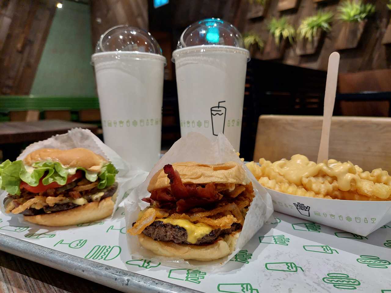 A selection of food from Shake Shack, displayed on a tray in a restaurant: a crispy shallot burger, a ShackMeister with bacon, Shack sauce, cheesy fries, two drinks