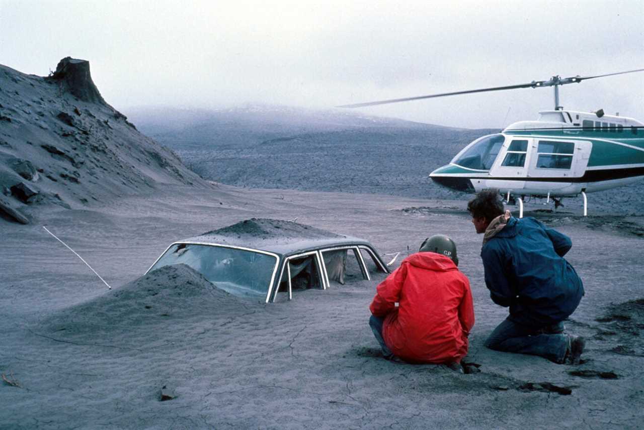 Two people kneel by a car covered in ash by a helicopter