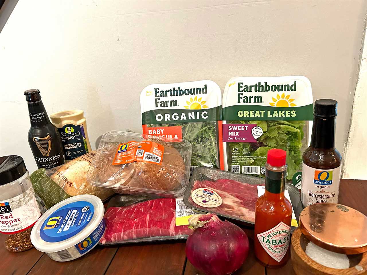 Ingredients on a table: a bottle of Guinness, crushed red pepper, blue-cheese crumbles, bread, portobello mushrooms, steak, a red onion, containers of sweet mix and baby arugula, Tabasco, Worcestershire sauce, and salt.