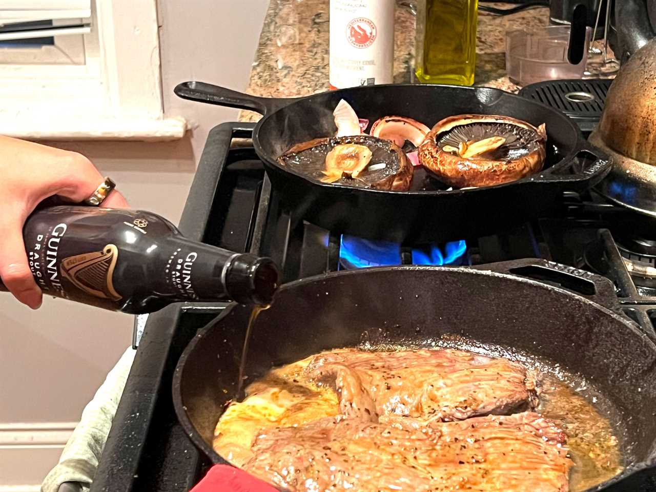 A hand pours Guinness beer into the skillet with the steak while the mushrooms and onions continue to cook on another burner.