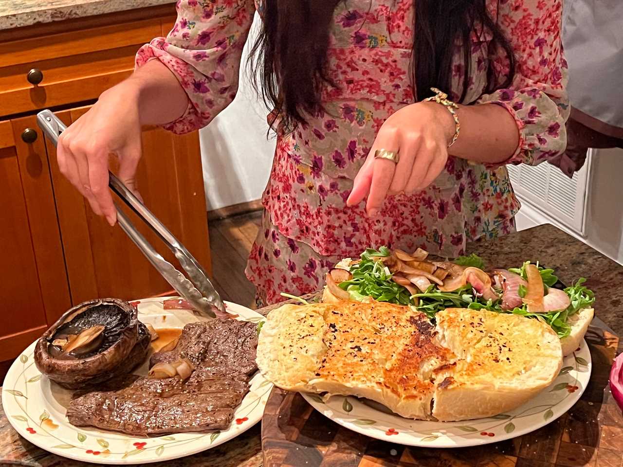 Lara uses tongs to pick up the steak on a plate. On another plate, the bread is laid out with the arugula and onions.