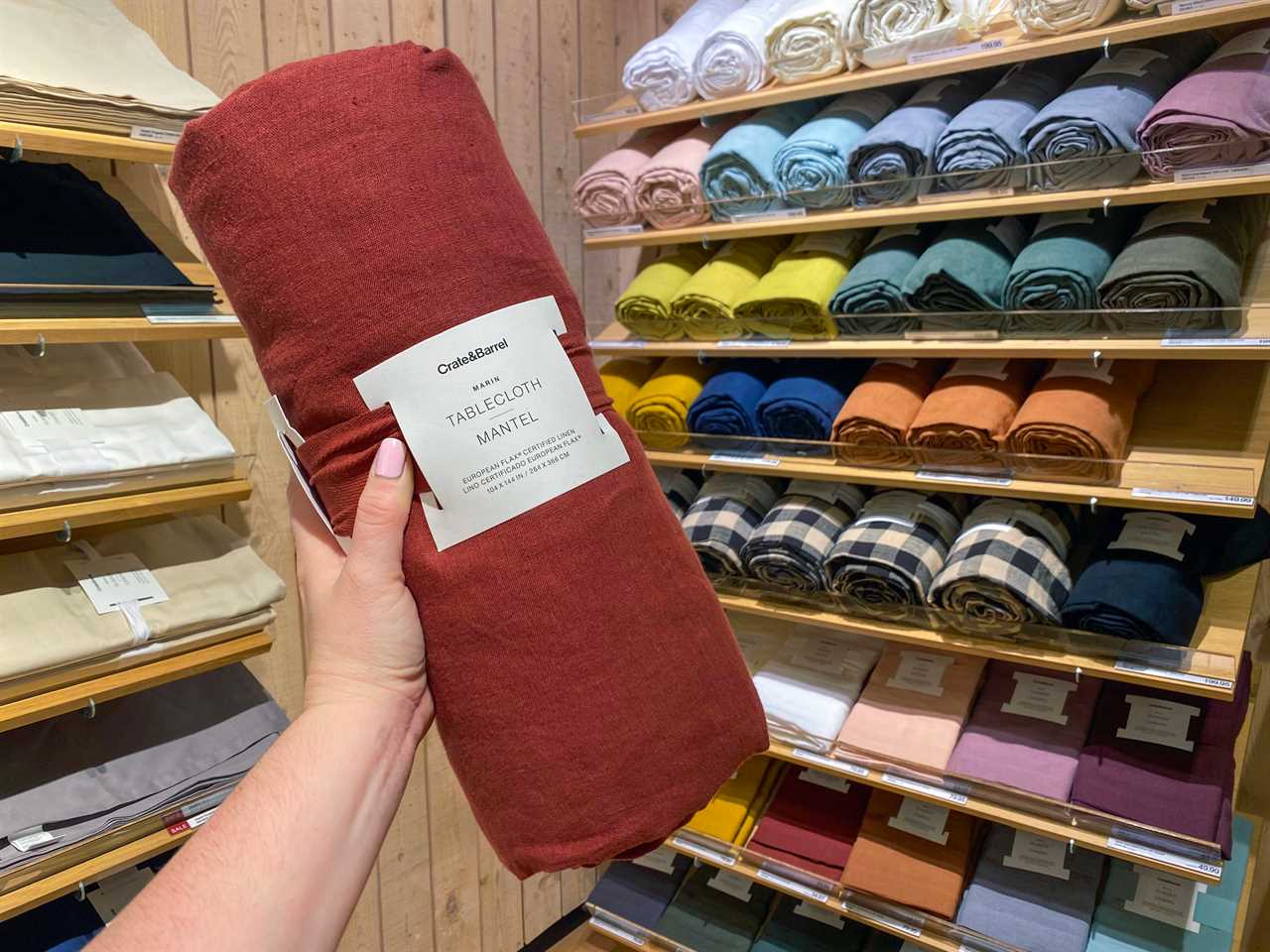 A hand holds a red rolled-up tablecloth in front of several rolled-up tablecloths in various colors on wooden shelves at Crate and Barrel