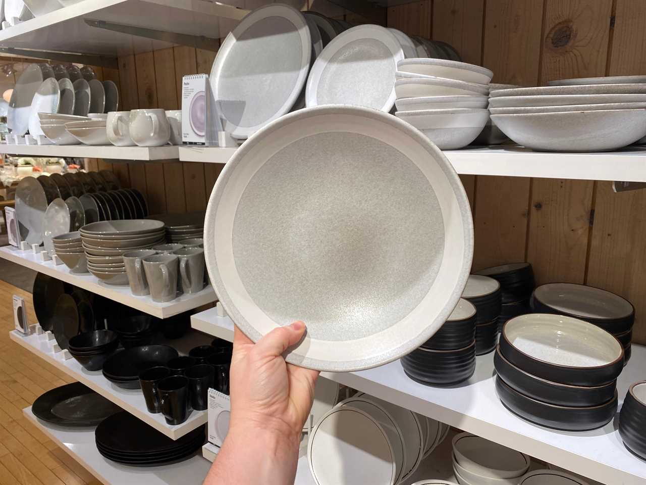 A hand holds a gray bowl with a white stripe along the rim in front of shelves of gray and black dish sets