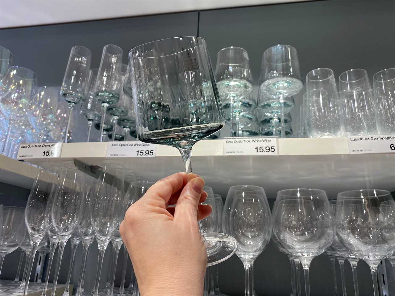 A hand holds a clear-glass drinking glass in front of shelves of several wine glasses. The glass in the foreground has a long stem and rectangular shape