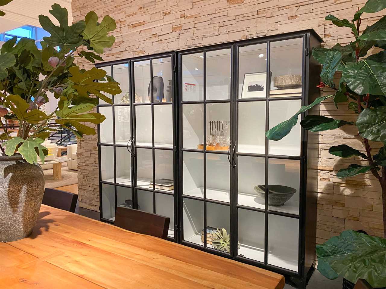 Metal cabinets with bowls, glasses, and decor in it against a stone wall  next to potted faux plants