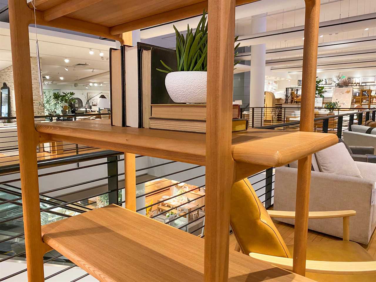 A wooden pass-through bookshelf, which has no back, with books and a faux plant with a white pot on top, at Crate and Barrel