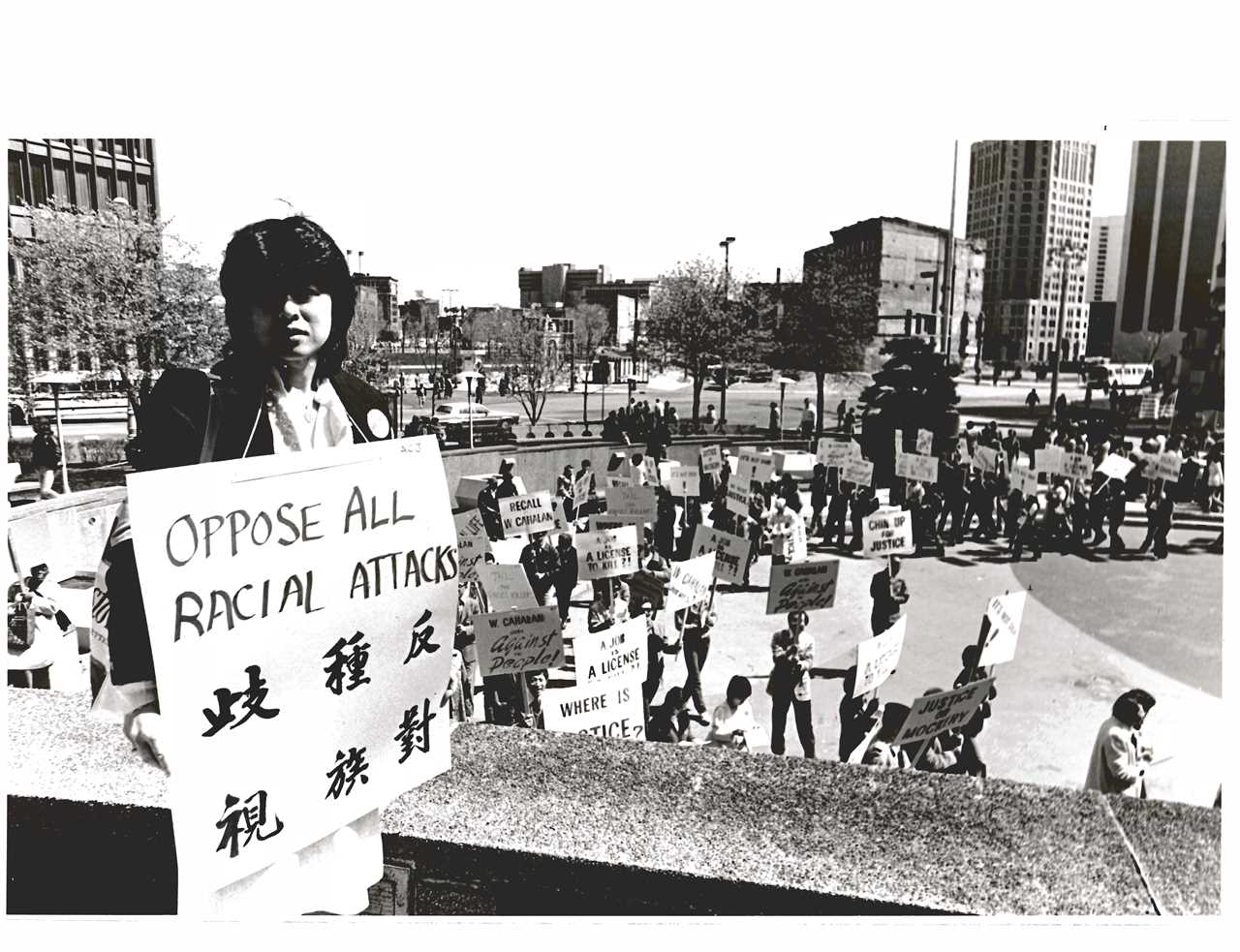 Helen Zia holding "Oppose All Racial Attacks" sign overlooking rally for justice for Vincent Chin.