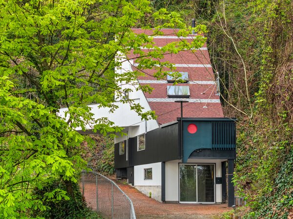 The home sits at the end of a long driveway, surrounded by tall trees and thriving greenery.