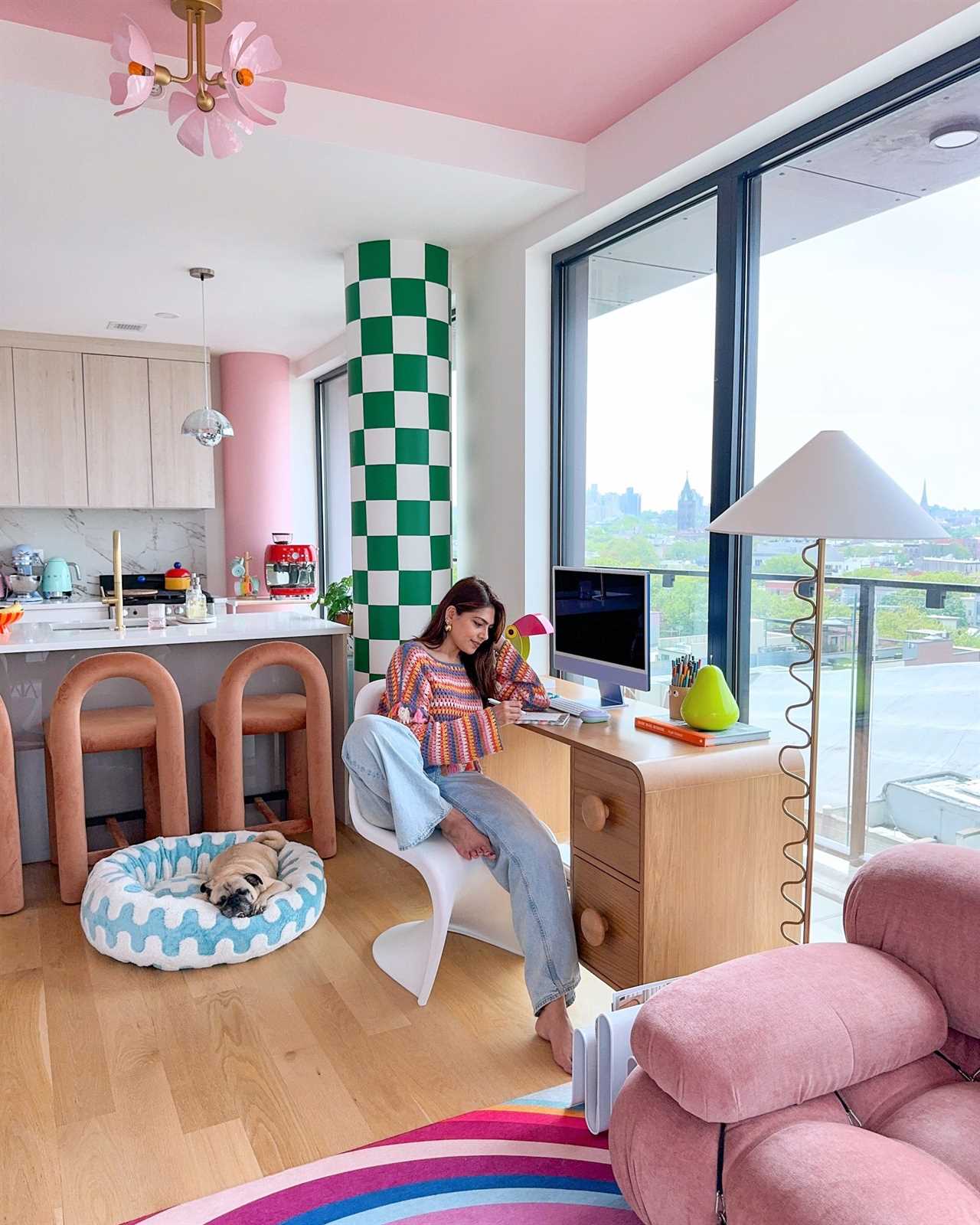 A woman sits at a desk in front of a window. The ceiling is pink, and her pug sits in a colorful dog bed.