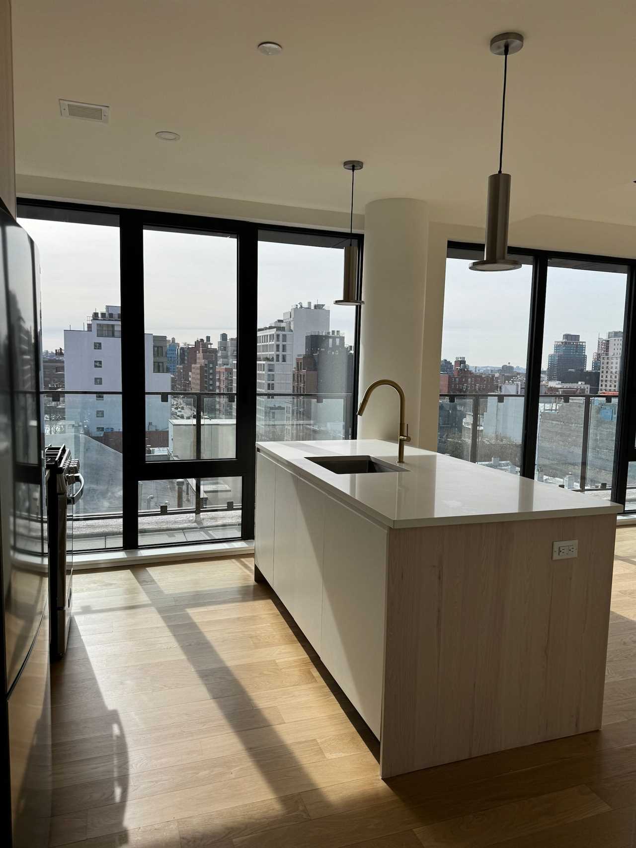 An empty kitchen in an apartment. The walls feature large windows and there's a large island in the center of the room.