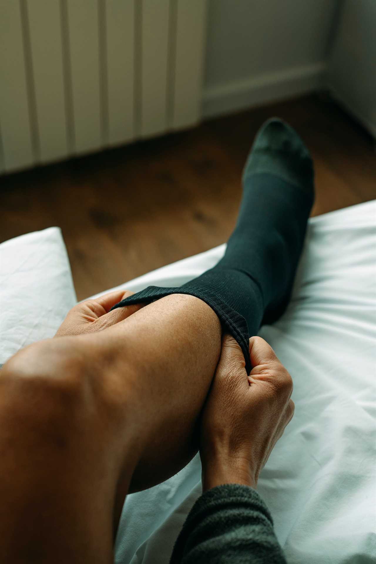 A man puts on black compression socks on a bed.