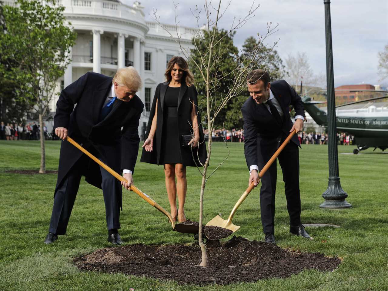 Donald Trump and Emmanuel Macron with shovels of dirt around a sapling. Melania Trump stands behind them watching.