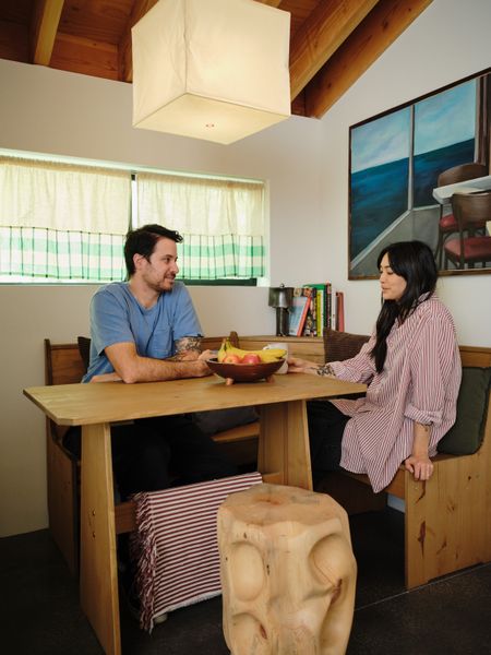 Rami Jrade and Hannah Go relax in the dining nook Hannah created in a corner of the open-plan living-dining room-kitchen. She found the bench and table online and the Akari pendant light from the Noguchi Museum website, and she made the cafe curtains with remnants from Ikea draperies. The stool was handcrafted by architect Hunter Knight, who designed and built the ADU. The painting is vintage.