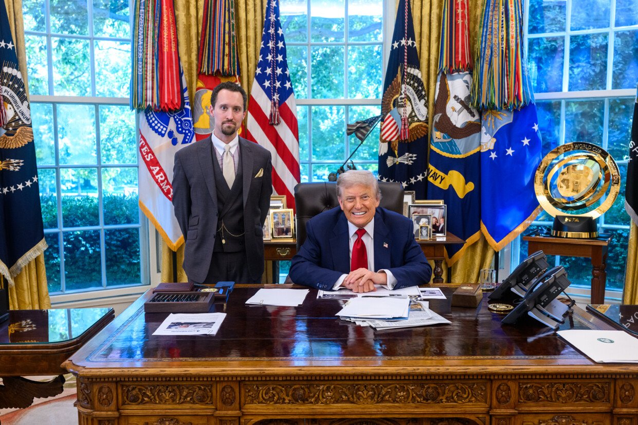 Donald Trump sitting at the Resolute Desk in the Oval Office with an aide standing beside him, surrounded by flags and official decor.