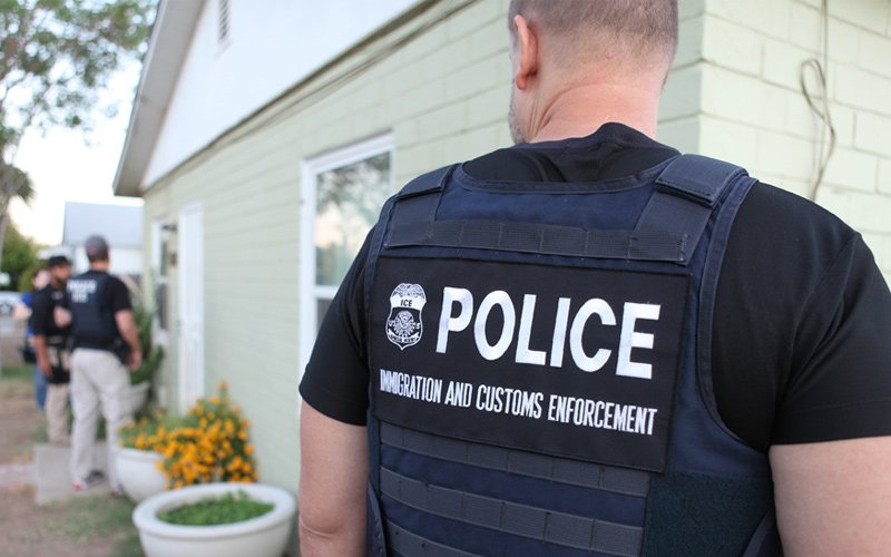 ICE officer wearing a tactical vest approaches a residence during an immigration enforcement operation.