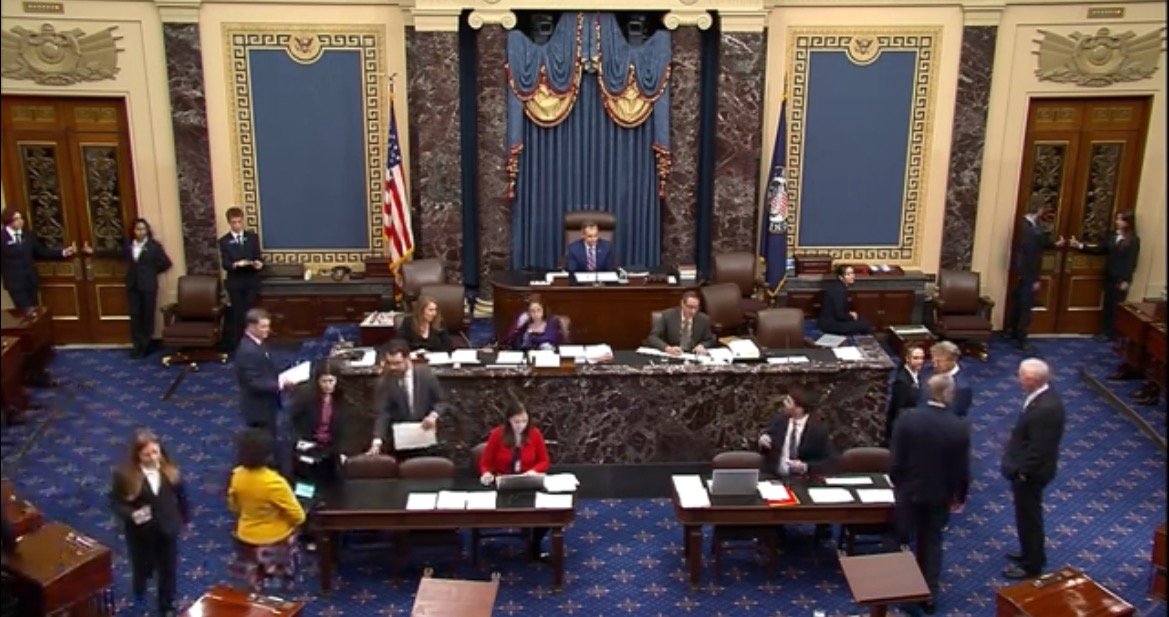 U.S. Senate chamber with senators and staff engaged in discussions, featuring the presiding officer's desk and American flags in the background.