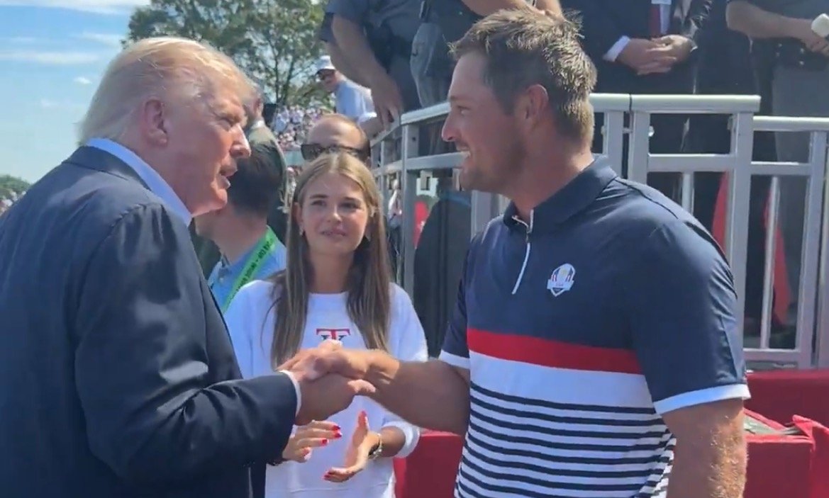 Donald Trump shakes hands with a golfer at a public event, while a young woman watches in the background on a sunny day.