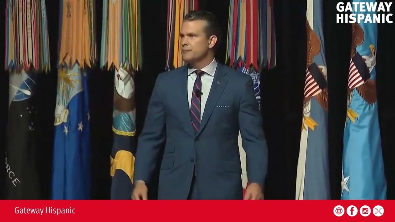 Speaker in a blue suit presents on stage with military flags in the background at a Gateway Hispanic event.